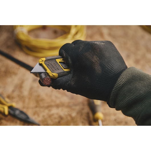 Photo of a STANLEY STHT10432 utility knife in a gloved hand cutting a cable on a work surface.