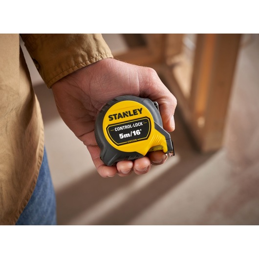 Photo of a STANLEY STHT37246-0 tape measure, CONTROL-LOCK 5m/16', held in a hand, construction background.