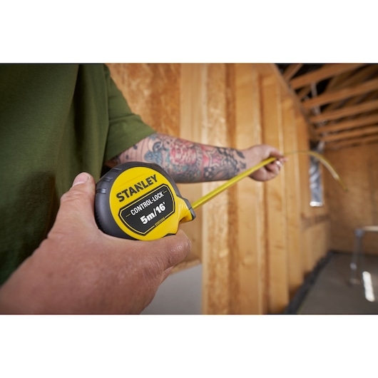 Stanley STHT37246-0 tape measure held in hand, measuring distance in unfinished wood-frame room.