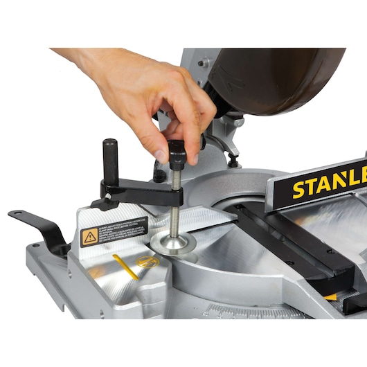 Photo of STANLEY STSM1525 miter saw, close-up side angle, hand adjusting the clamp on the metal base.
